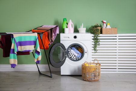 Modern washing machine, detergents, houseplant and clothes near light green wall indoorsの写真素材