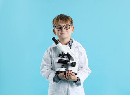 Cute little boy in laboratory coat and glasses with microscope on light blue background. Child and scienceの写真素材