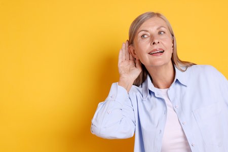 Senior woman showing hand to ear gesture on orange background, space for text
