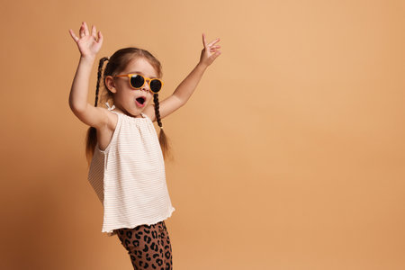 Stylish little girl with pigtails in sunglasses having fun on beige background. Space for text