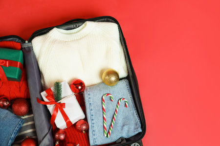 Christmas vacation. Open suitcase with clothes, gift box, baubles and candy canes on red background, top view. Space for textの写真素材