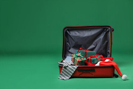 Christmas vacation. Open suitcase with Santa hat, clothes and gift boxes on green background. Space for textの写真素材