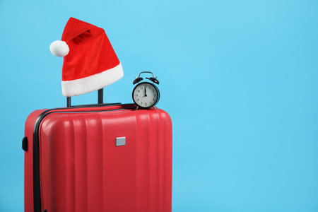 Christmas vacation. Suitcase with red Santa hat and alarm clock on light blue background. Space for textの写真素材