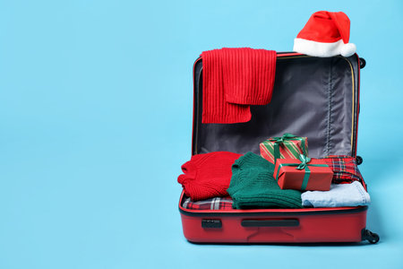 Christmas vacation. Open suitcase with Santa hat, clothes and gift boxes on light blue background. Space for textの写真素材