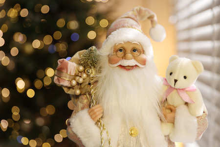 Santa Claus figure with sack of gifts and toy bear against blurred lights indoors, closeup with space for text. Merry Christmas and Happy New Yearの写真素材