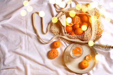 Fresh tangerines, glass with sparkling wine and fir branch on white bedsheet, flat lay. Bokeh lights