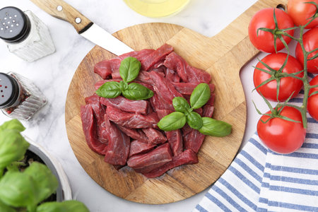 Pieces of raw beef meat, basil and ingredients on white marble table, flat lay