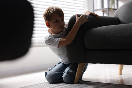 Child in danger. Frightened little boy hiding behind armchair at homeの写真素材