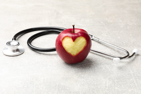 Ripe apple with carved heart and stethoscope on gray textured table, closeup