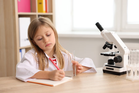 Cute little girl with microscope and test tube writing at desk indoors. Child and scienceの写真素材
