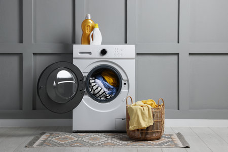 Modern washing machine, detergents and clothes near gray wall indoors