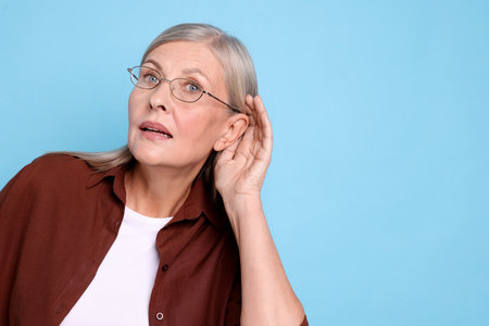 Senior woman showing hand to ear gesture on light blue background, space for text