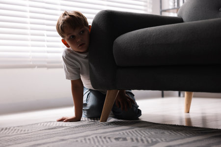 Child in danger. Frightened little boy hiding behind armchair at homeの写真素材