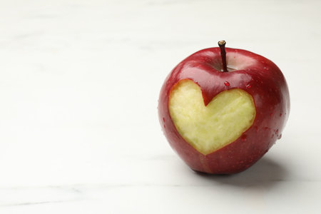 Ripe apple with carved heart on white marble table, closeup. Space for text