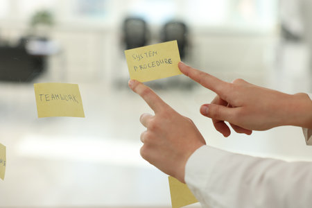 Woman sticking note with text System Procedure on glass board in office, closeup