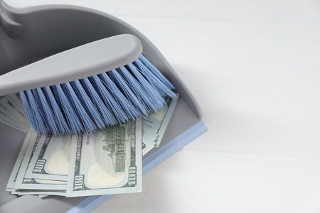 Dustpan with broom and dollar banknotes on white wooden surface, closeup. Space for textの写真素材