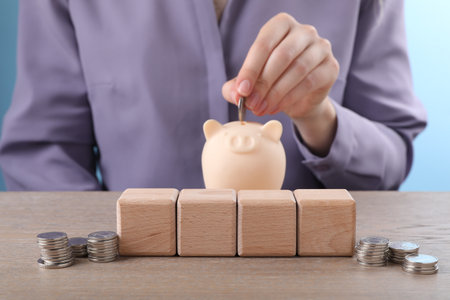 Empty cubes and stacked coins on table, selective focus. Woman putting coin into piggy bank, closeupの写真素材