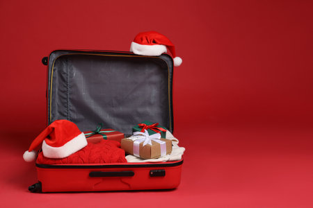 Christmas vacation. Open suitcase with Santa hats, clothes and gift boxes on red background. Space for textの写真素材