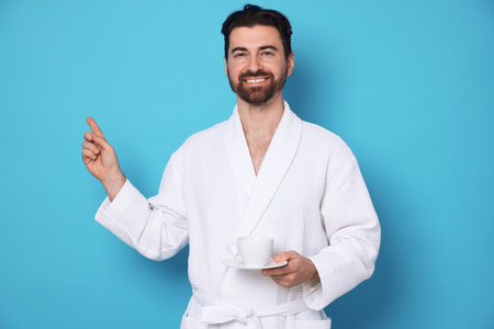 Man in bathrobe with cup of coffee pointing at something on light blue backgroundの写真素材