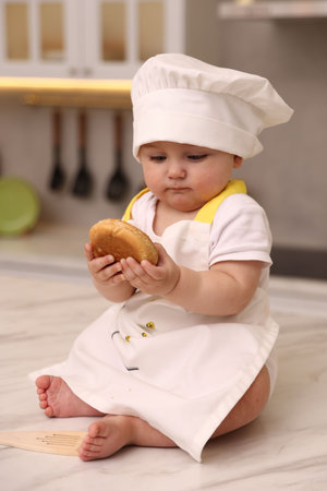 Cute baby with bun wearing chef uniform at light marble table in kitchenの写真素材
