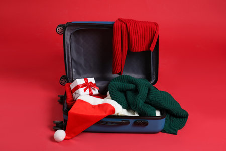 Christmas vacation. Open suitcase with Santa hat, clothes and gift box on red backgroundの写真素材