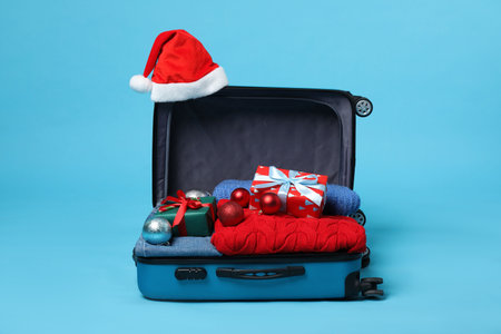 Christmas vacation. Open suitcase with Santa hat, clothes, gift boxes and baubles on light blue backgroundの写真素材
