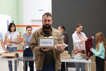 Volunteers helping homeless people in shelter, focus on man with box of donations