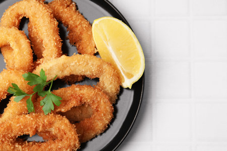 Deep fried squid rings with parsley and lemon slice on white tiled table, closeup. Space for text