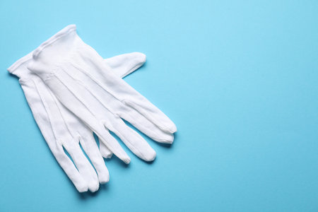 Pair of white cloth gloves on light blue background, top view. Space for text