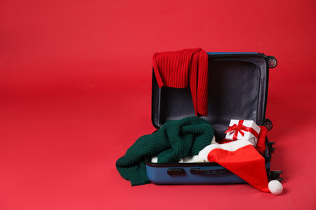 Christmas vacation. Open suitcase with Santa hat, clothes and gift box on red background. Space for textの写真素材