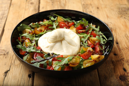Delicious burrata cheese with arugula, tomatoes and dressing on wooden table, closeup