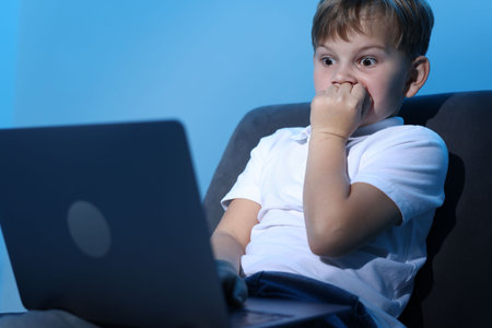 Cyber danger. Frightened little boy using laptop on blue background