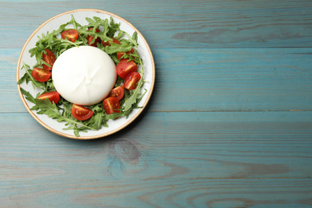 Delicious burrata cheese with tomatoes and arugula on light blue wooden table, top view. Space for...