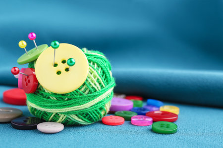 Ball of bright knitting yarn with buttons and pins on blue fabric, closeup. Space for textの写真素材