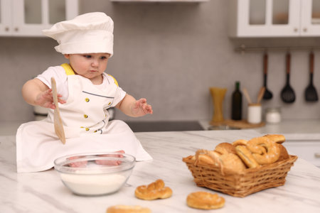 Cute baby with spatula wearing chef uniform at light marble table in kitchen