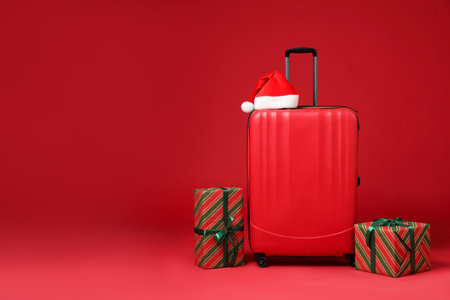 Christmas vacation. Stylish suitcase, Santa hat and gift boxes on red background. Space for textの写真素材