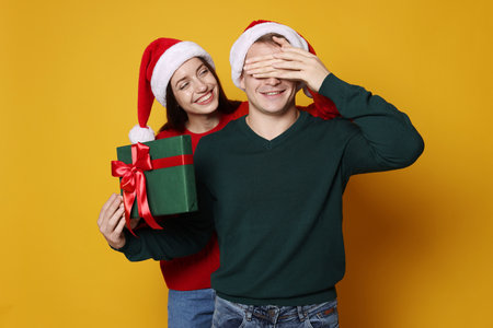 Smiling woman surprising her boyfriend with Christmas gift on yellow background. Happy couple in Santa hatsの写真素材
