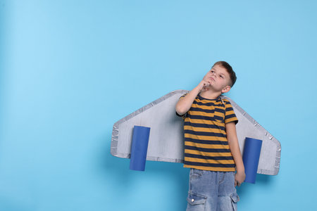 Little boy with cardboard plane wings on light blue background. Space for text