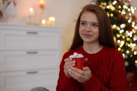 Beautiful teenage girl holding cup of hot drink with marshmallows against blurred lights at home, space for text. Christmas atmosphereの写真素材