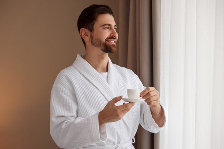Man in bathrobe with cup of coffee near window indoors. Space for textの写真素材