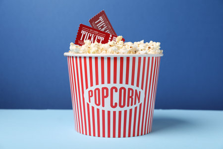Tasty popcorn in paper cup and movie tickets on table against blue background, closeupの写真素材