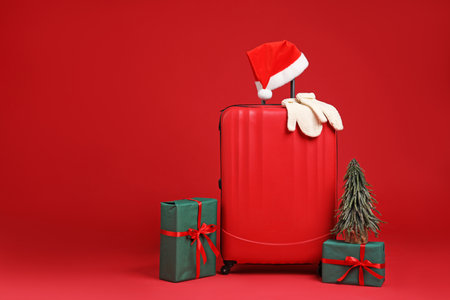 Christmas vacation. Stylish suitcase with Santa hat, mittens, gift boxes and Christmas tree on red background. Space for textの写真素材