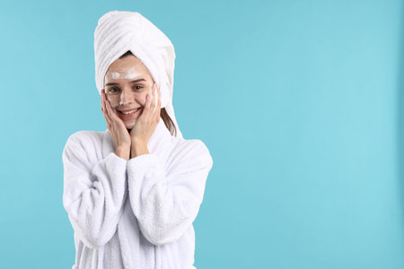 Beautiful woman with hair wrapped in towel applying cosmetic product on face against light blue background. Space for textの写真素材