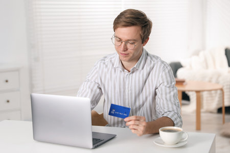 Young man making online payment with laptop and credit card at white table indoorsの写真素材