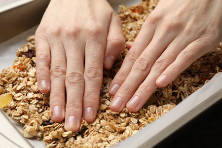 Making granola. Woman putting oats flakes with dried fruits onto tray at gray table, closeupの写真素材