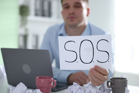 Man with laptop and sign SOS at table in office, selective focus