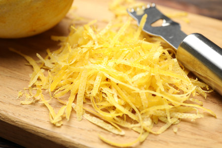 Fresh lemon zest and zester tool on wooden board, closeup