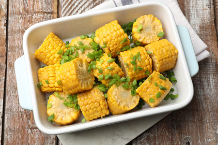 Pieces of boiled corncobs with green onion in baking dish on wooden table, top view