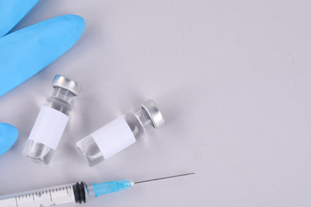 Vaccine in glass vials, medical gloves and syringe on gray background, flat lay. Space for text