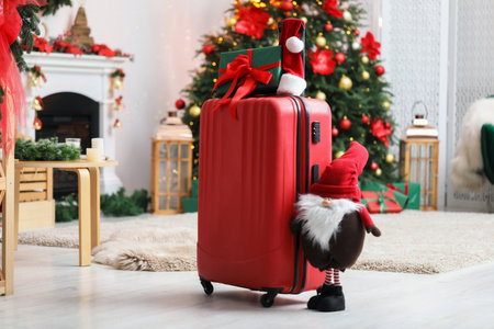 Suitcase with gift, Santa hat and decorative dwarf at home against blurred lights. Christmas vacationの写真素材